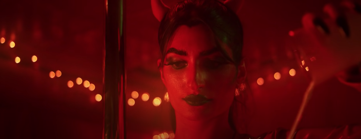 A devil like vixen with a red face, horns, and black lipstick stands beside a pole dancing pole with her eyes cast down in an authoritative manner.